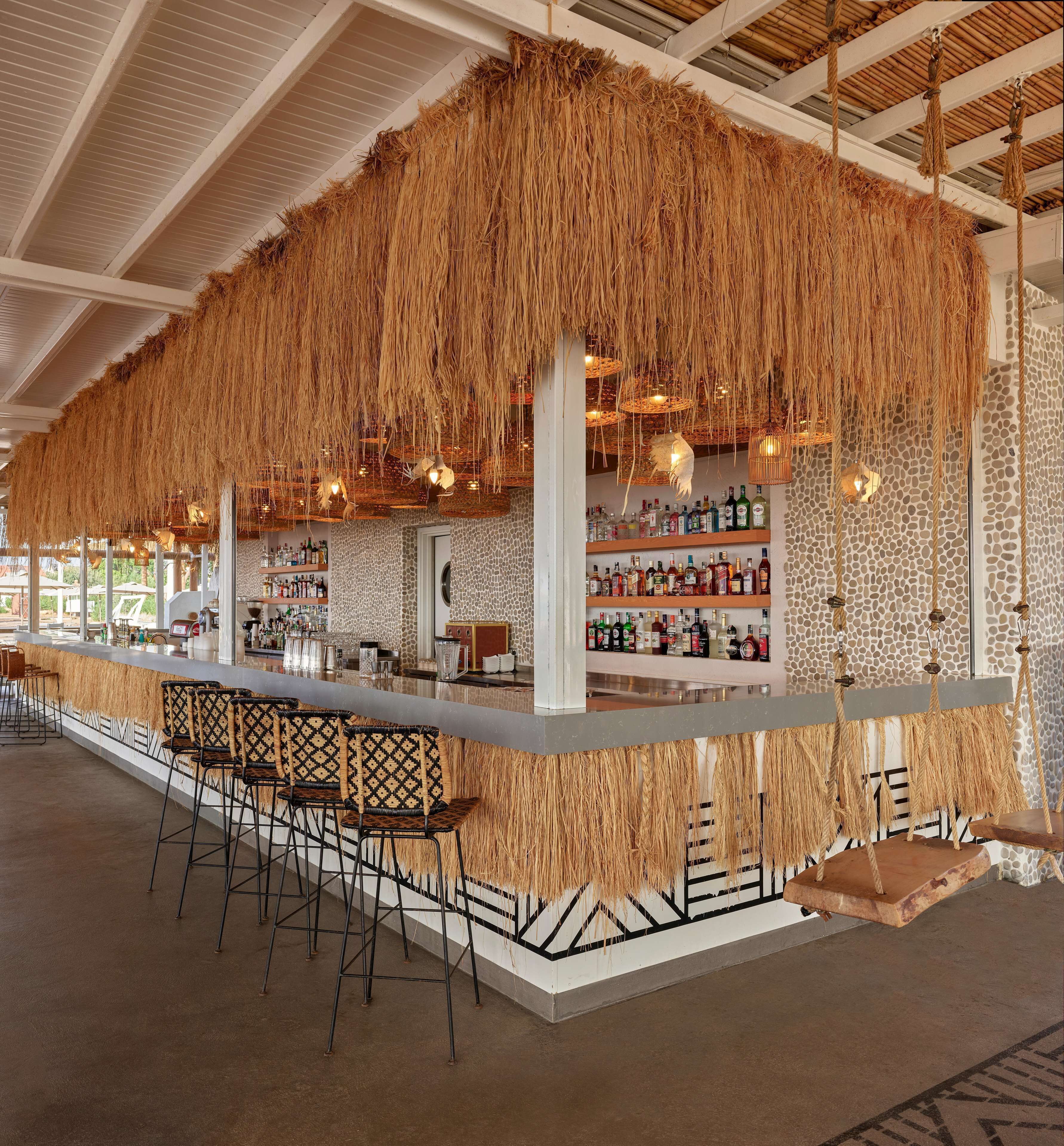 Zona de bar con techo de paja, taburetes y una variedad de botellas en estanterías.