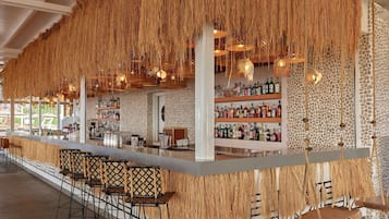 Zona de bar con techo de paja, taburetes y una variedad de botellas en estanterías.