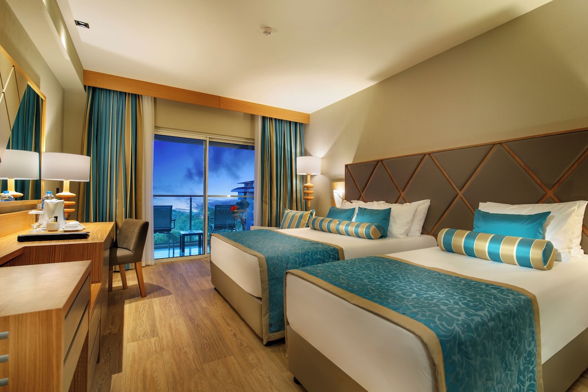 A hotel room with two beds, a desk, a chair, a TV, and a balcony with a view.