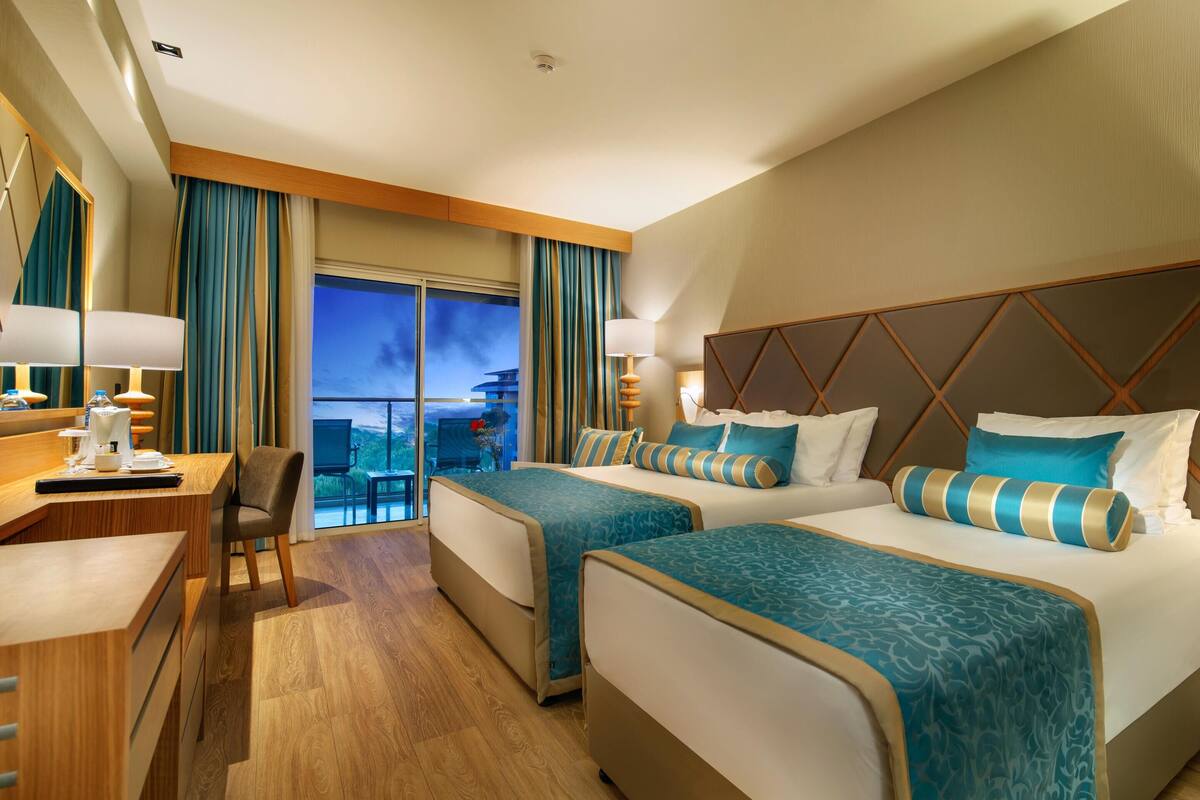 A hotel room with two beds, a desk, a chair, a TV, and a balcony with a view of a building.