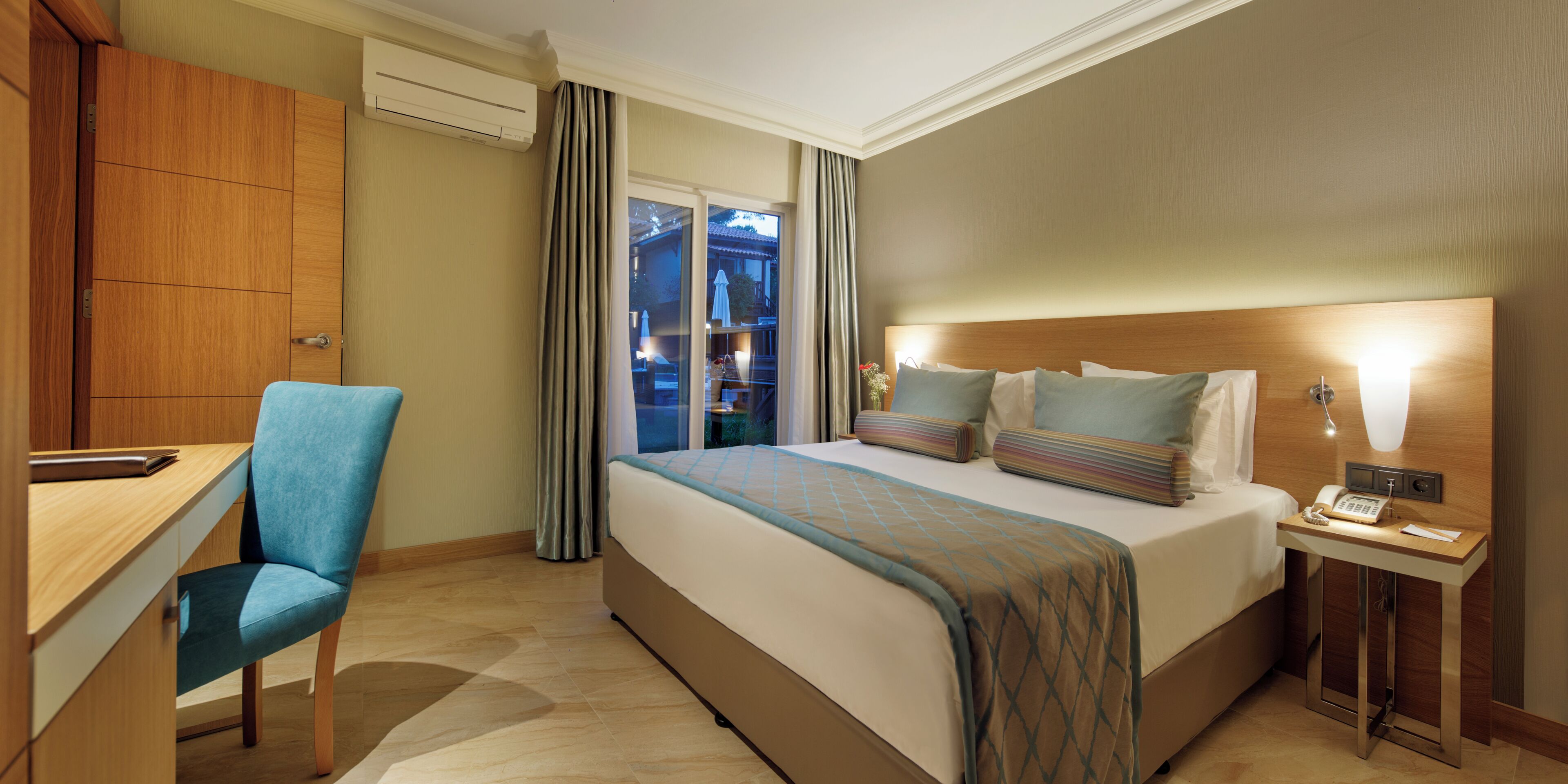 A hotel room with a bed, a desk, a chair, a bedside table with a phone, and a view of the outside.
