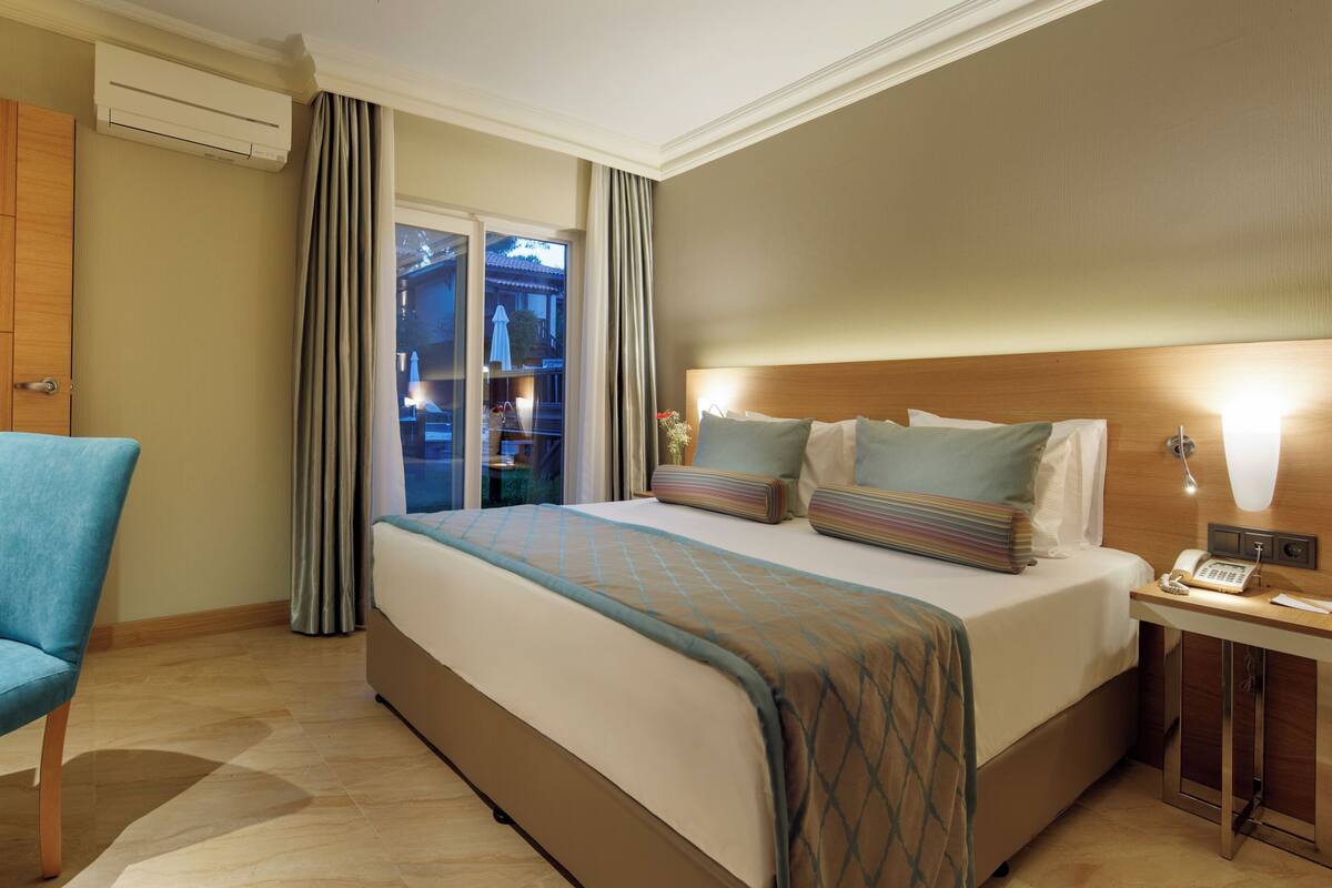 A hotel room with a bed, a desk, a chair, a bedside table with a phone, and a view of the outside.