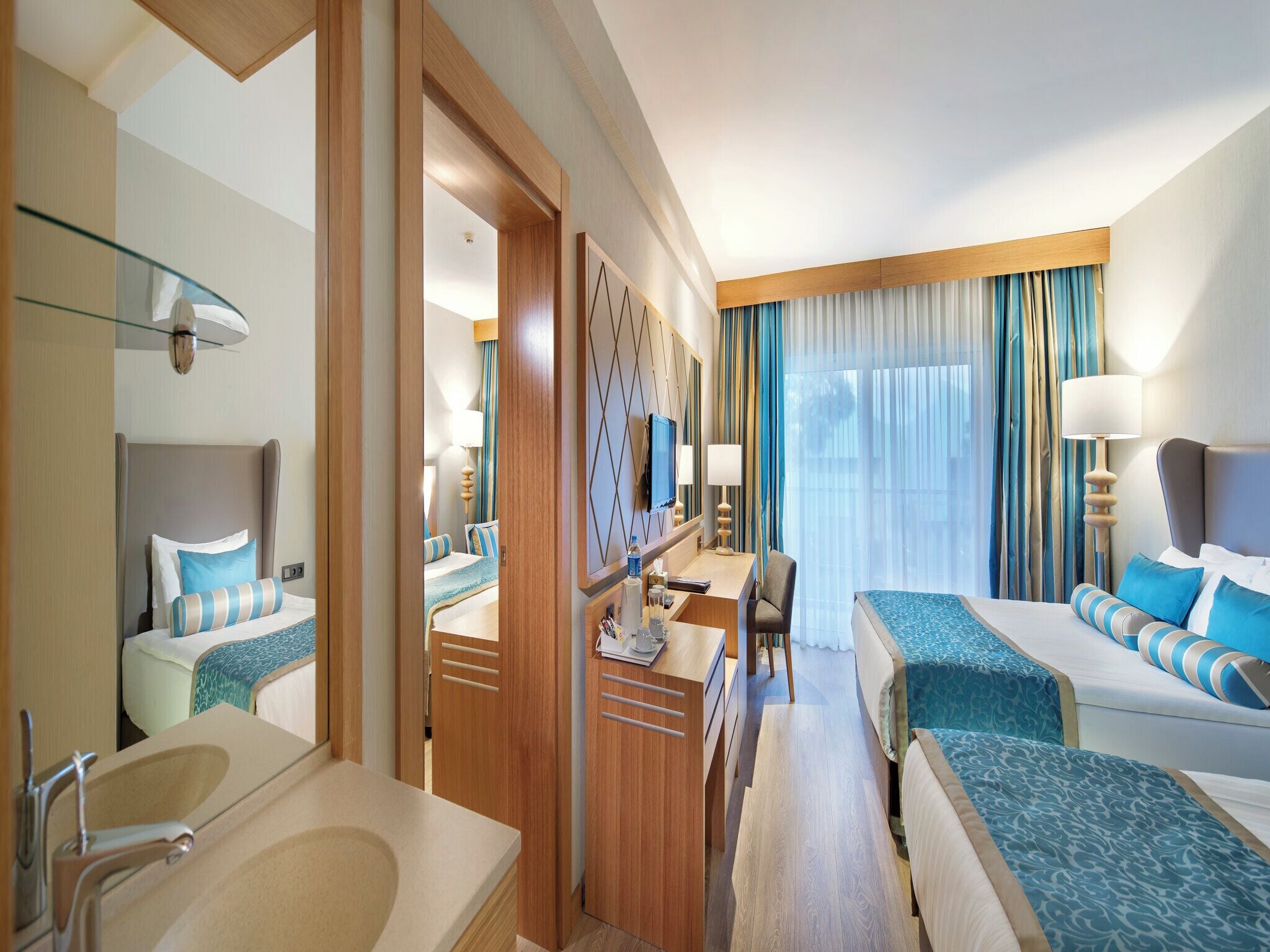 A hotel room with a large bed, a desk, a chair, a bathroom with a sink and mirror, and a large window with curtains.