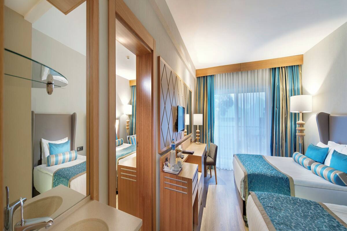 A hotel room with a large bed, a desk, a chair, a bathroom with a sink and mirror, and a large window with curtains.
