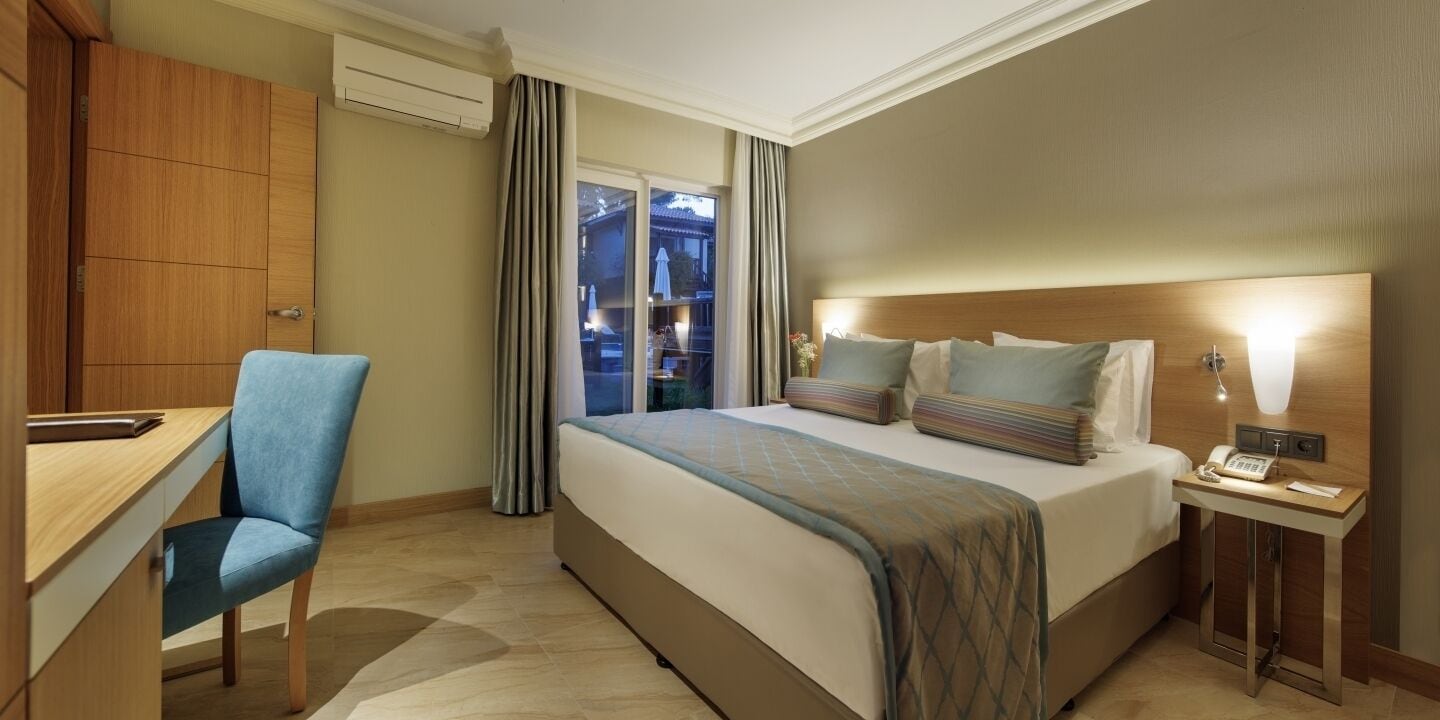 A hotel room with a bed, a desk, a chair, a bedside table with a lamp, and a view through a window.