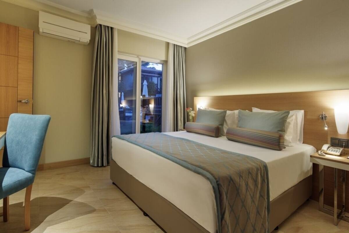 A hotel room with a bed, a desk, a chair, a bedside table with a lamp, and a view of the outside through a window.