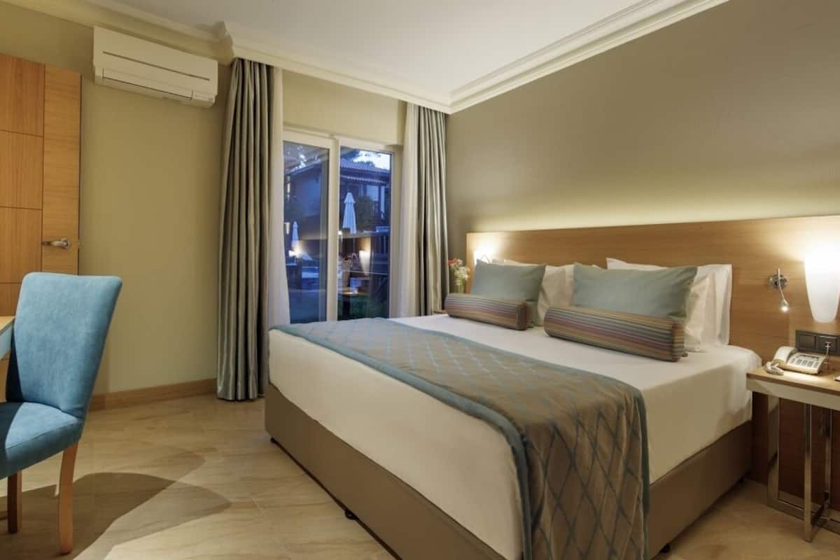 A hotel room with a bed, a desk, a chair, a bedside table with a lamp, and a view through a window.