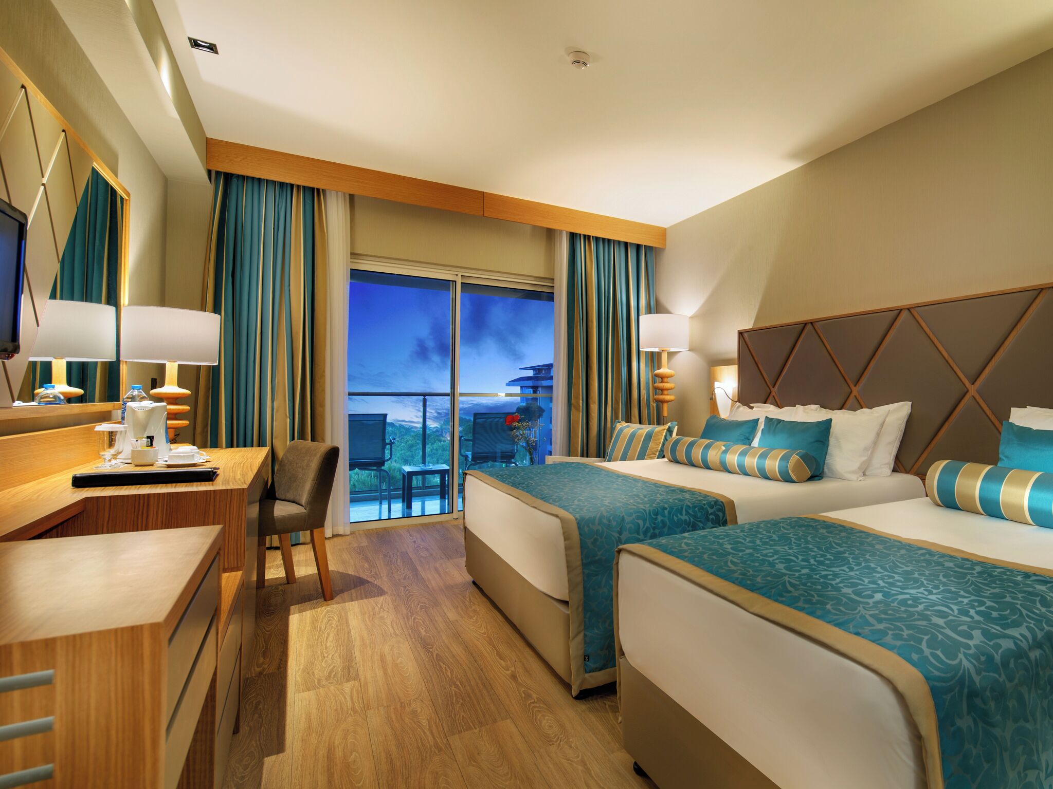 A hotel room with two beds, a desk, and a chair. There is a large window with curtains, a television, and a lamp.