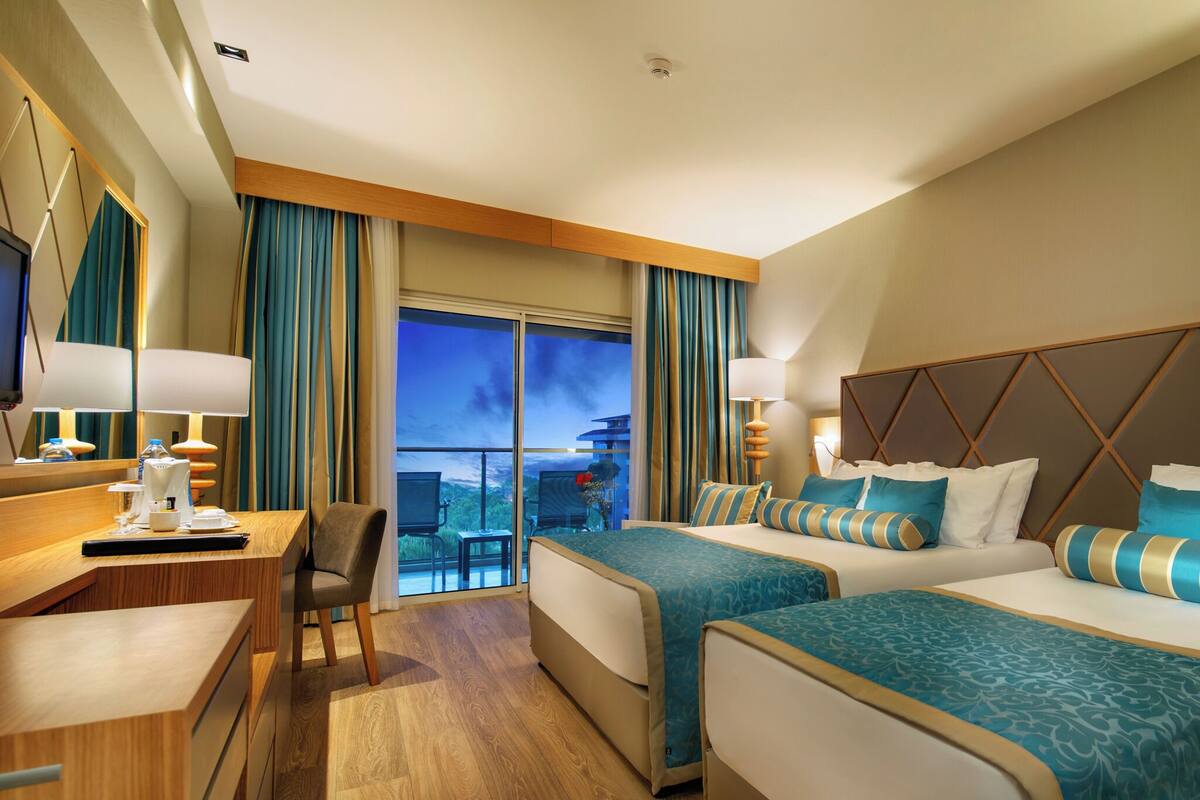 A hotel room with two beds, a desk, and a chair. There is a large window with curtains, a television, and a lamp.