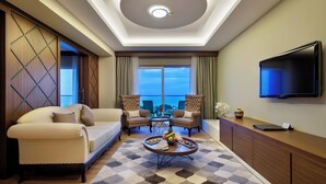 Ein modernes Hotelzimmer mit einer Couch, zwei Sesseln, einem Couchtisch mit Obst und Snacks, einem Flachbildfernseher und Blick auf die Stadt durch große Fenster.