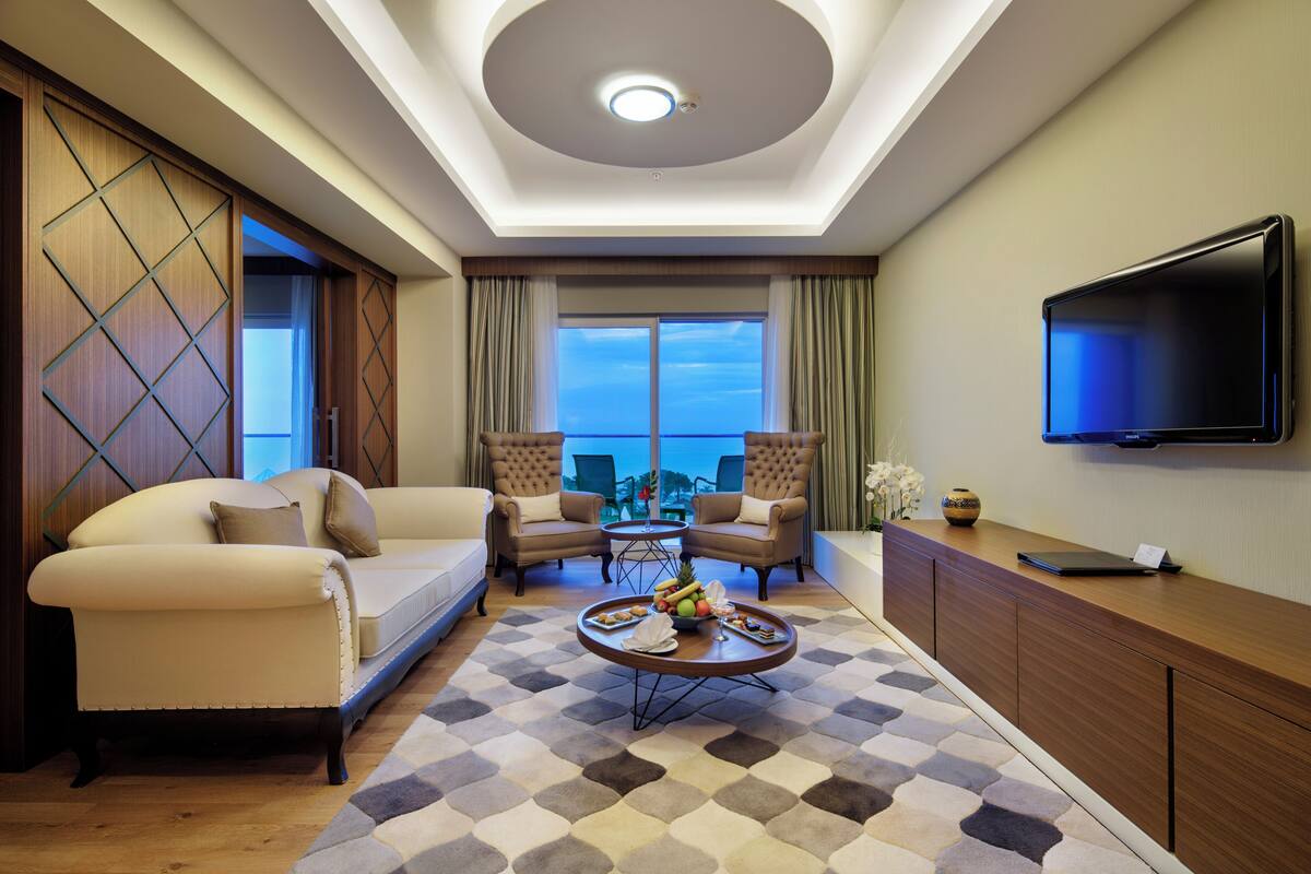 A modern hotel room with a sofa, two armchairs, a coffee table with fruit and snacks, a flat-screen TV, and a view of the city through large windows.