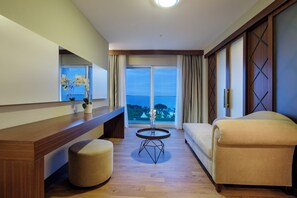 Ein modernes Hotelzimmer mit Holzboden, einem beigefarbenen Sofa, einem runden Tisch mit Blumenvase und einem großen Fenster mit Vorhängen.