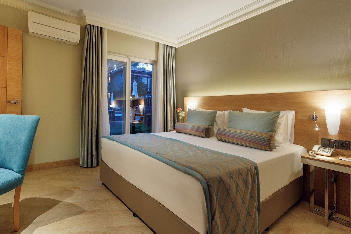 A hotel room with a bed, a desk, a chair, a bedside table with a phone, and a view of the outside.