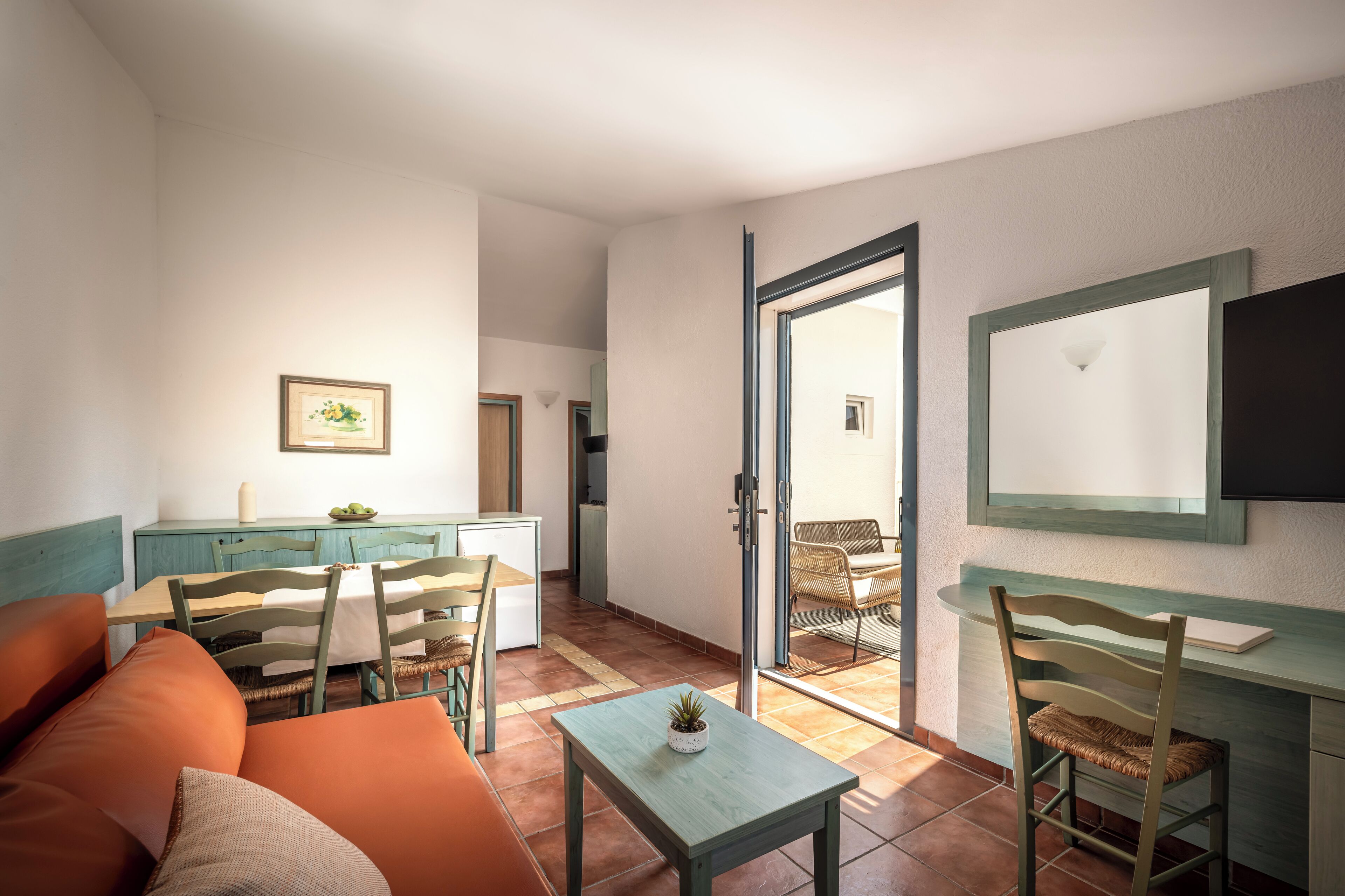 Un espace de vie compact comprenant un canapé, une table à manger, des chaises, une petite table avec une plante, un coin cuisine et une porte-fenêtre coulissante donnant sur un espace extérieur.