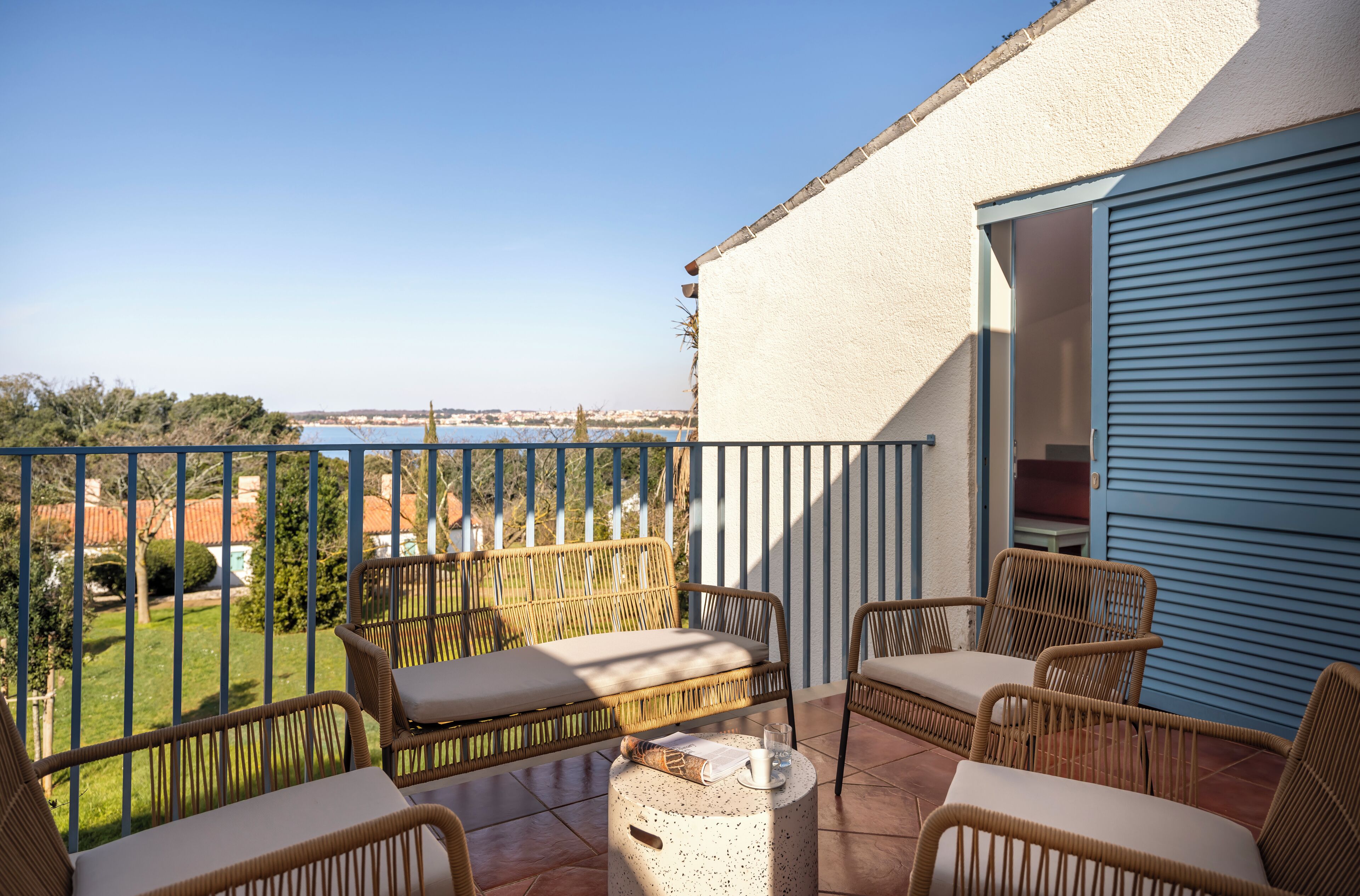 A balcony with wicker furniture, a view of the sea, and a building with a blue shutter.