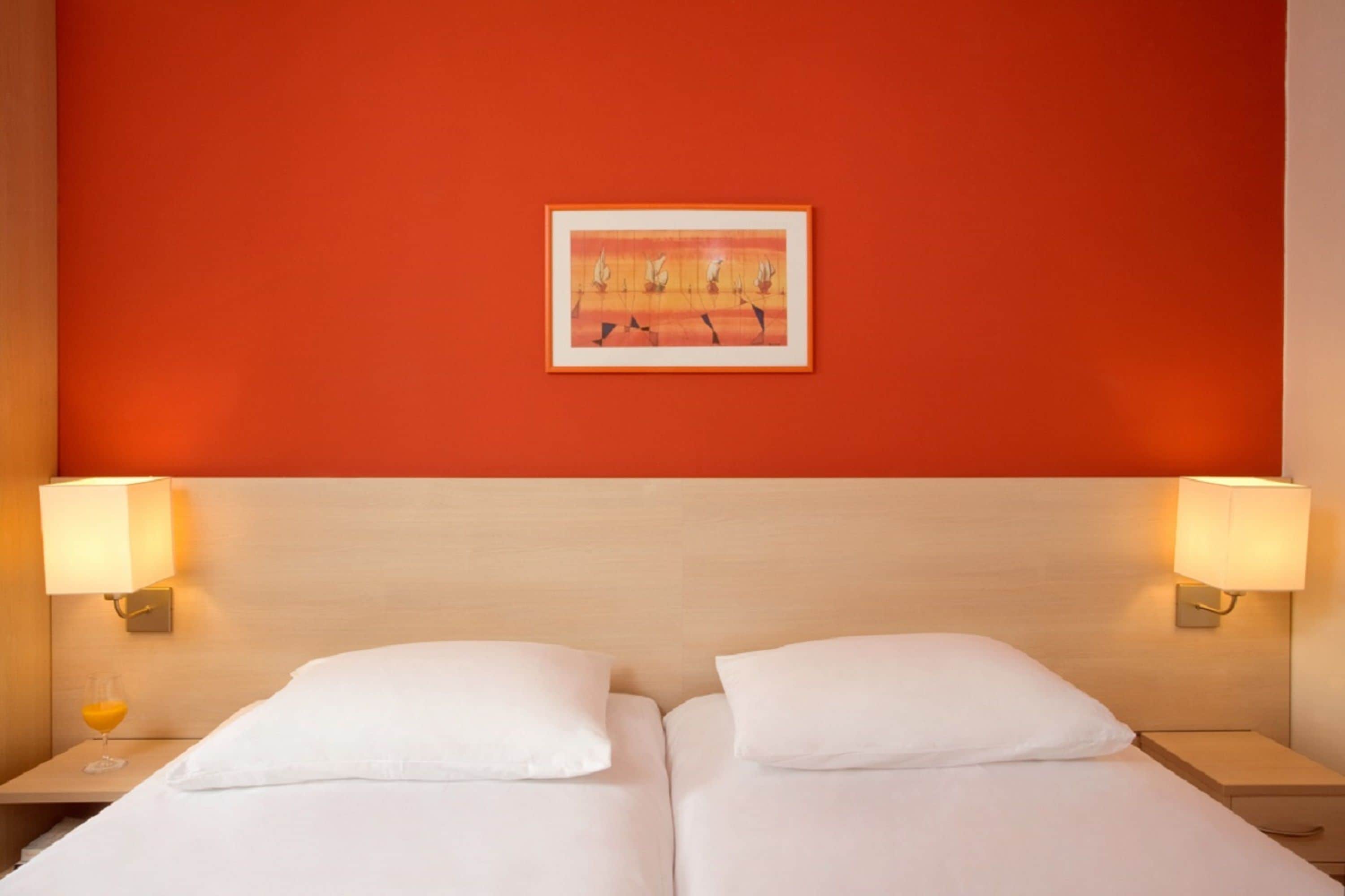 Ein Hotelzimmer mit zwei Betten, einer roten Wand und einem gerahmten Kunstwerk darüber.