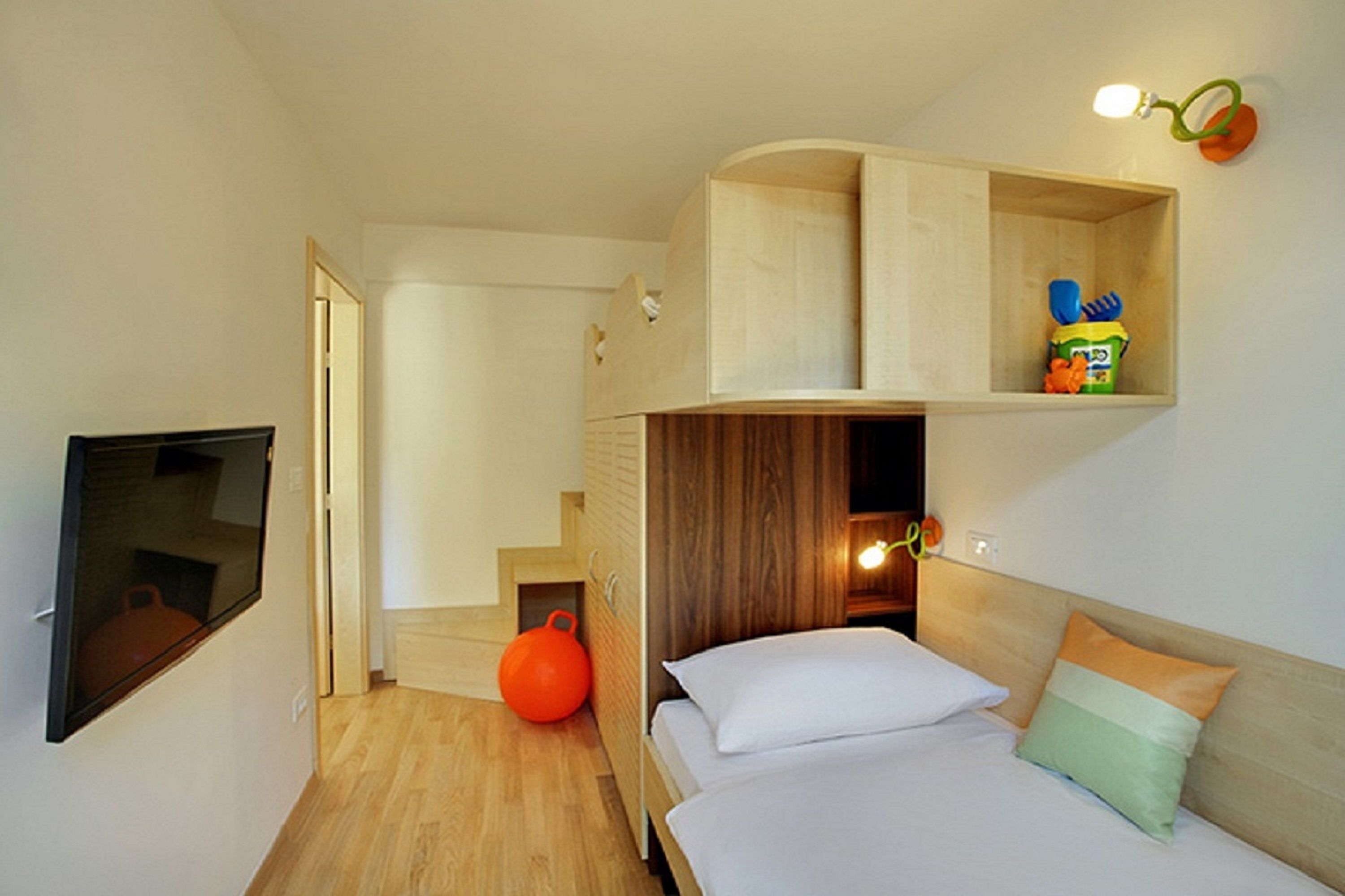 A compact hotel room with a bed, a TV, a wooden shelf with toiletries, and a small orange exercise ball.