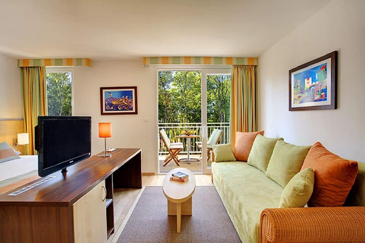 A hotel room with a bed, a TV, a desk, a sofa, a balcony with a table and chairs, and two paintings on the walls.