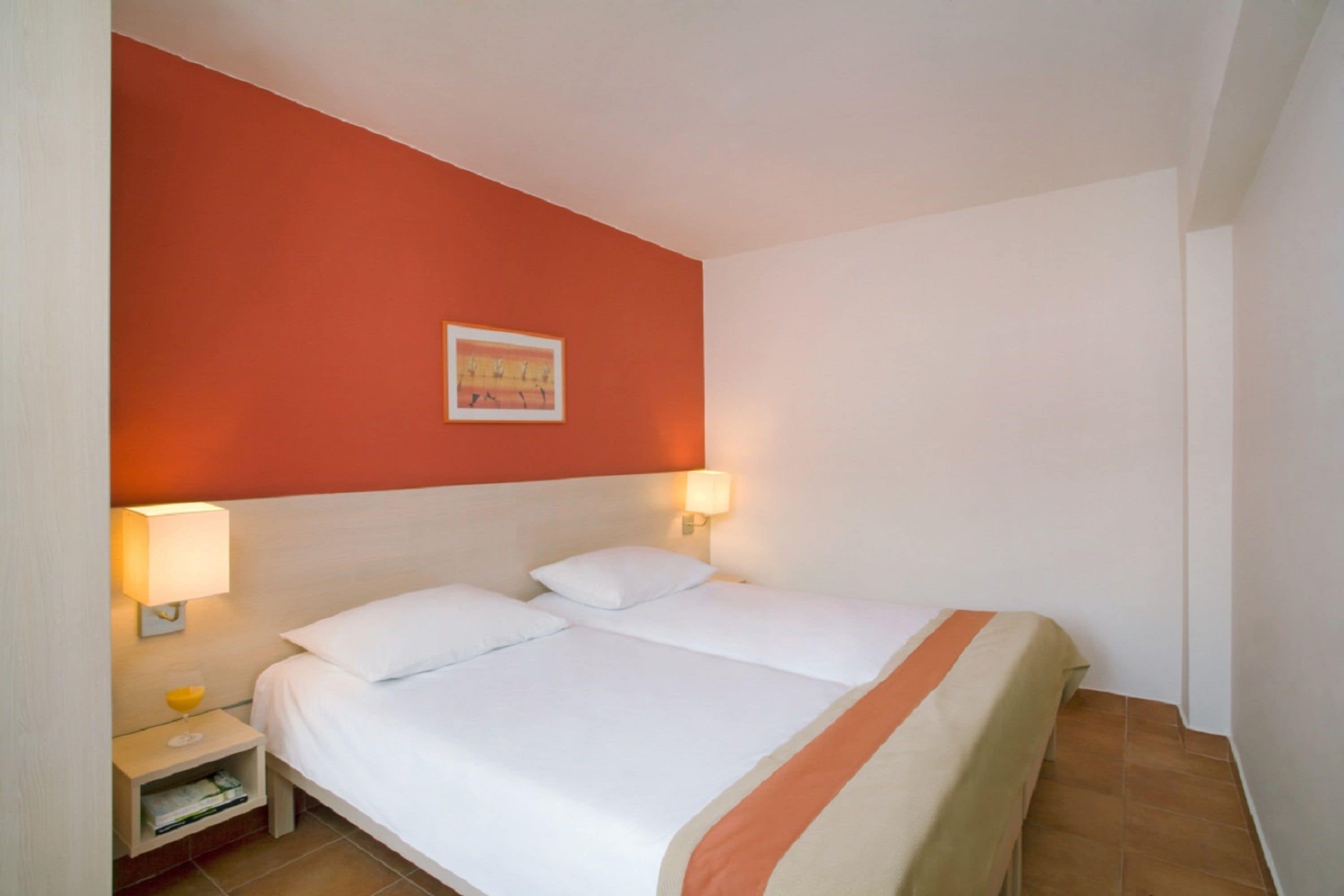 A hotel room with a bed, two bedside lamps, a nightstand with a glass of orange juice, and a framed picture on the wall.