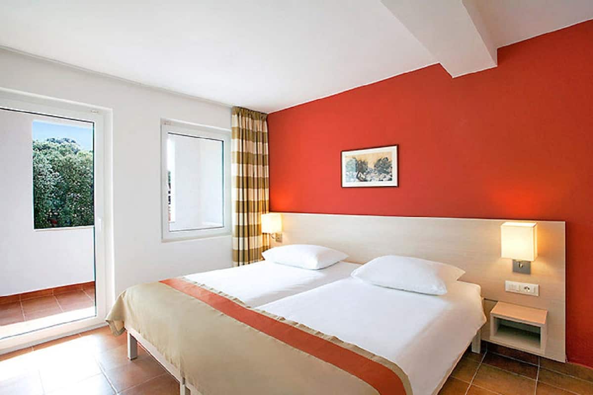 A hotel room with a large bed, two bedside tables, a window with curtains, and a red accent wall.