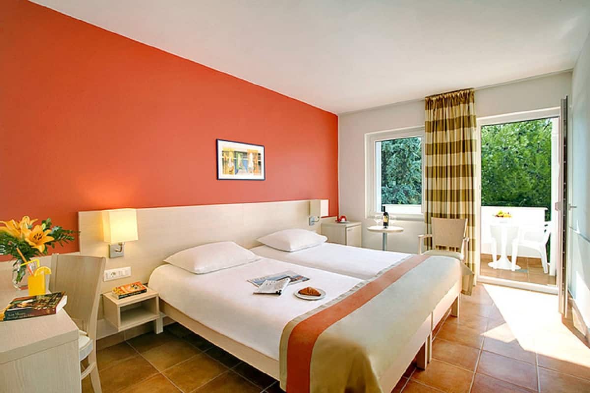 A hotel room with two beds, a window with curtains, a small table with a lamp, and a view of greenery outside.