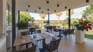 Un área de comedor al aire libre con varias mesas y sillas, rodeada de amplias ventanas que ofrecen vistas a palmeras y vegetación.