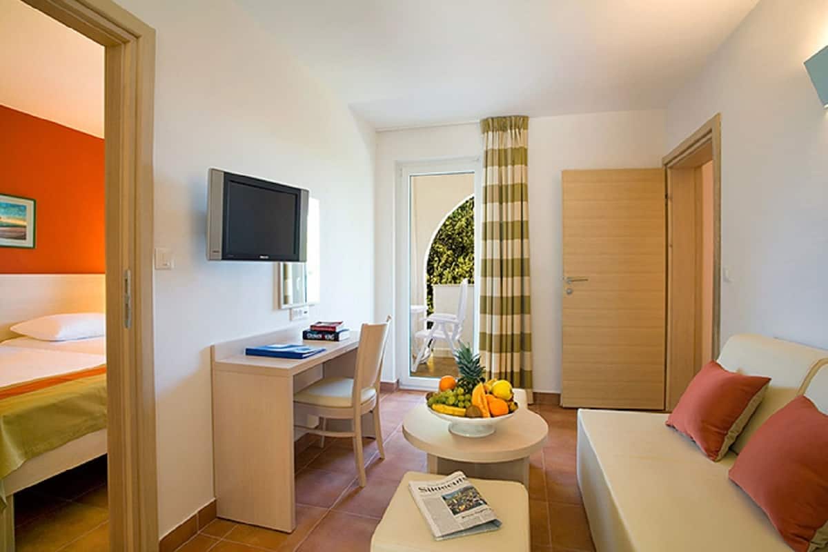 A compact hotel room with a bed, a desk, a television, a small table with fruit, and a sofa.