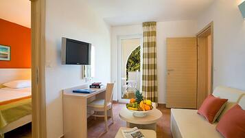 A compact hotel room with a bed, a desk, a television, a small table with fruit, and a sofa.