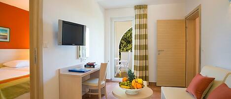 A compact hotel room with a bed, a desk, a television, a small table with fruit, and a sofa.