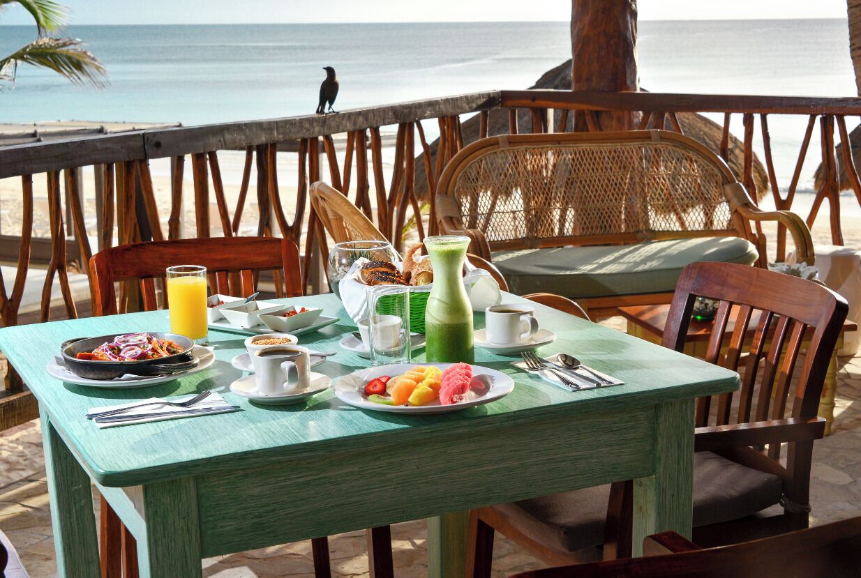 A table set with breakfast items, including a plate of fruit, a green drink, and a cup of coffee, overlooking a beach with a wooden railing and chairs.