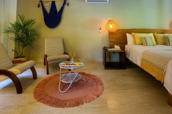 A hotel room with a bed, two chairs, a small table with snacks, a hanging hammock, a lamp, and a potted plant.