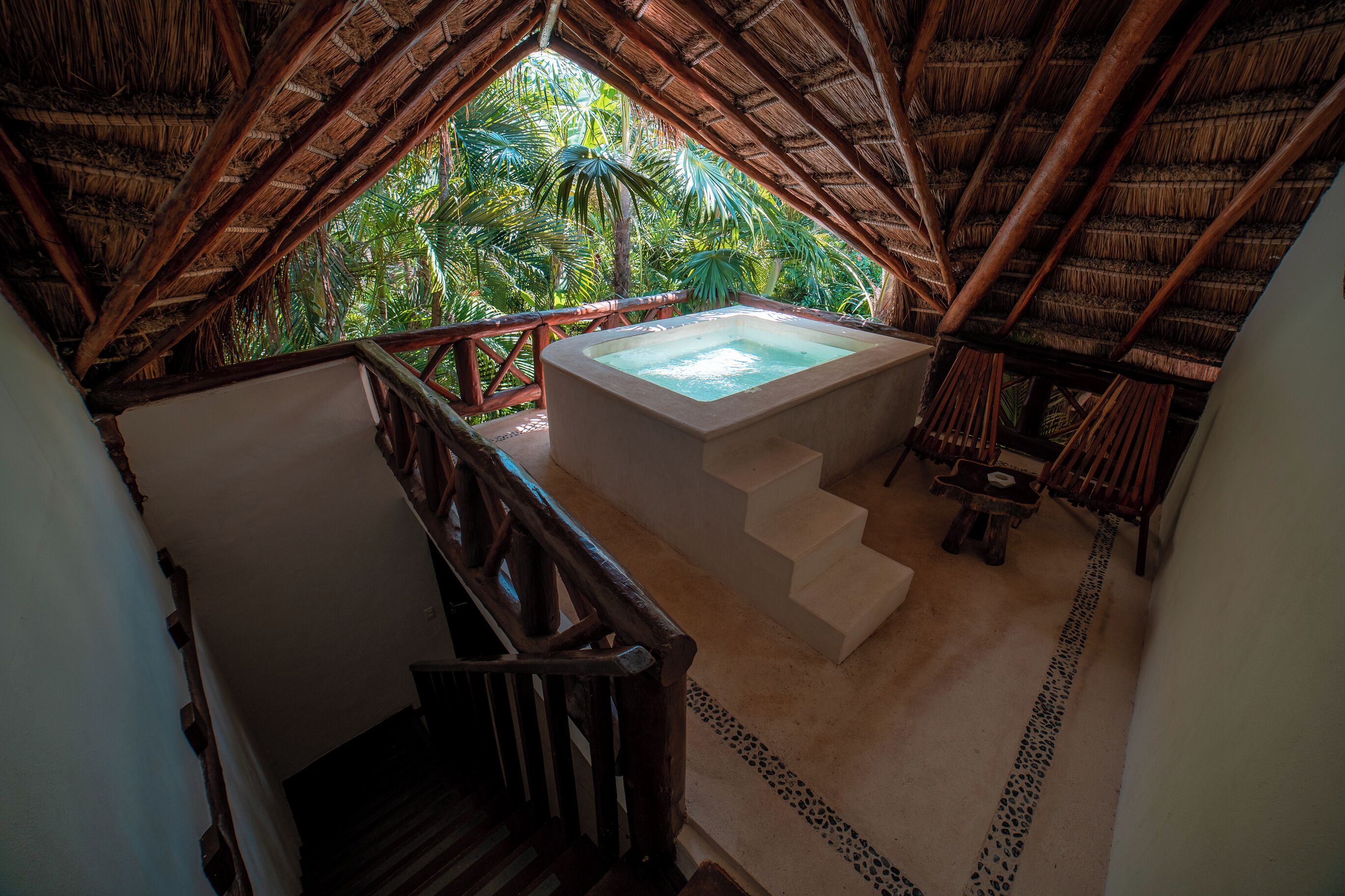 A thatched-roof room with a hot tub, a staircase, and a view of lush greenery.