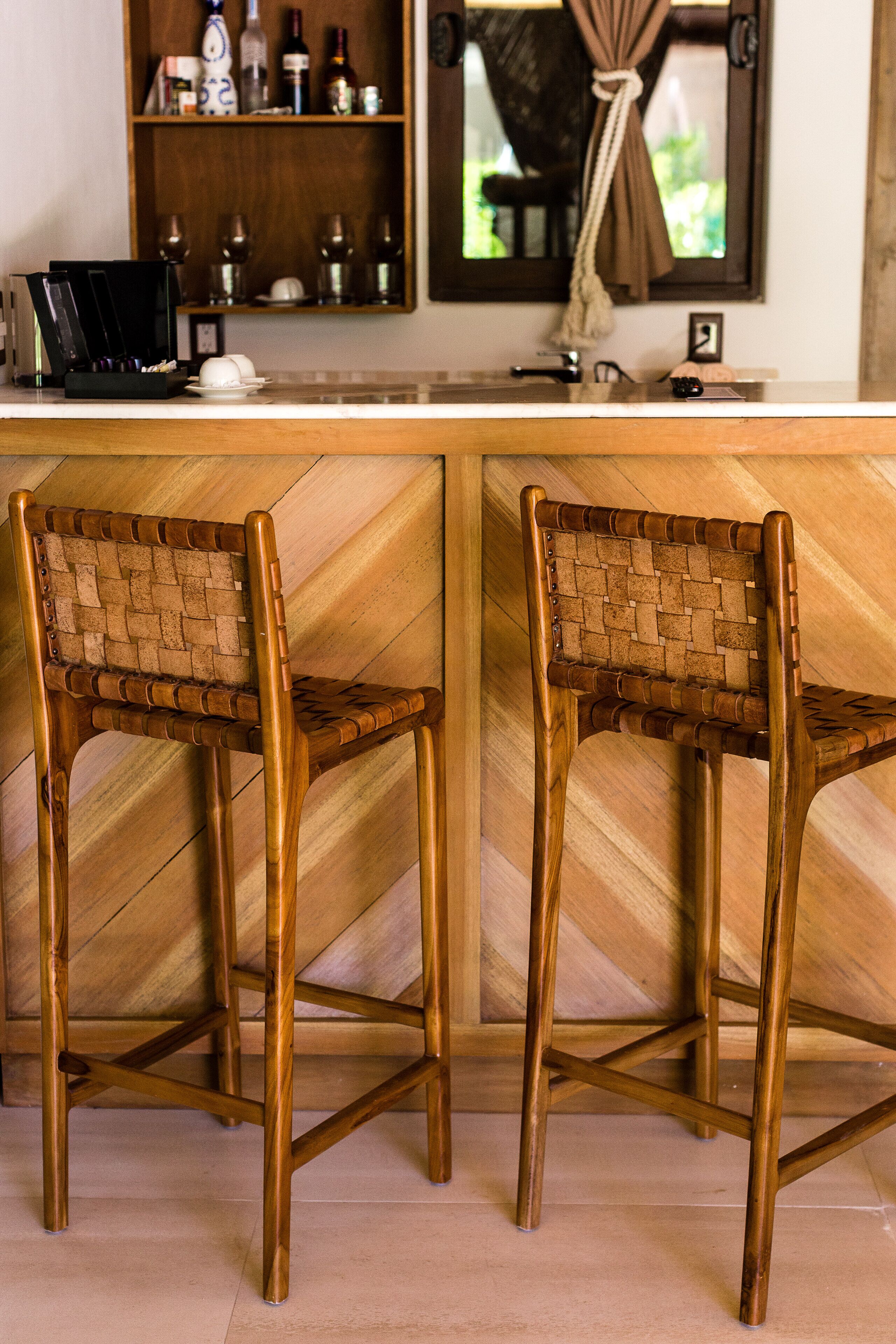 Zona de bar con dos sillas de madera con asientos y respaldos trenzados, una barra con un pequeño reloj y una estantería con botellas y vasos.