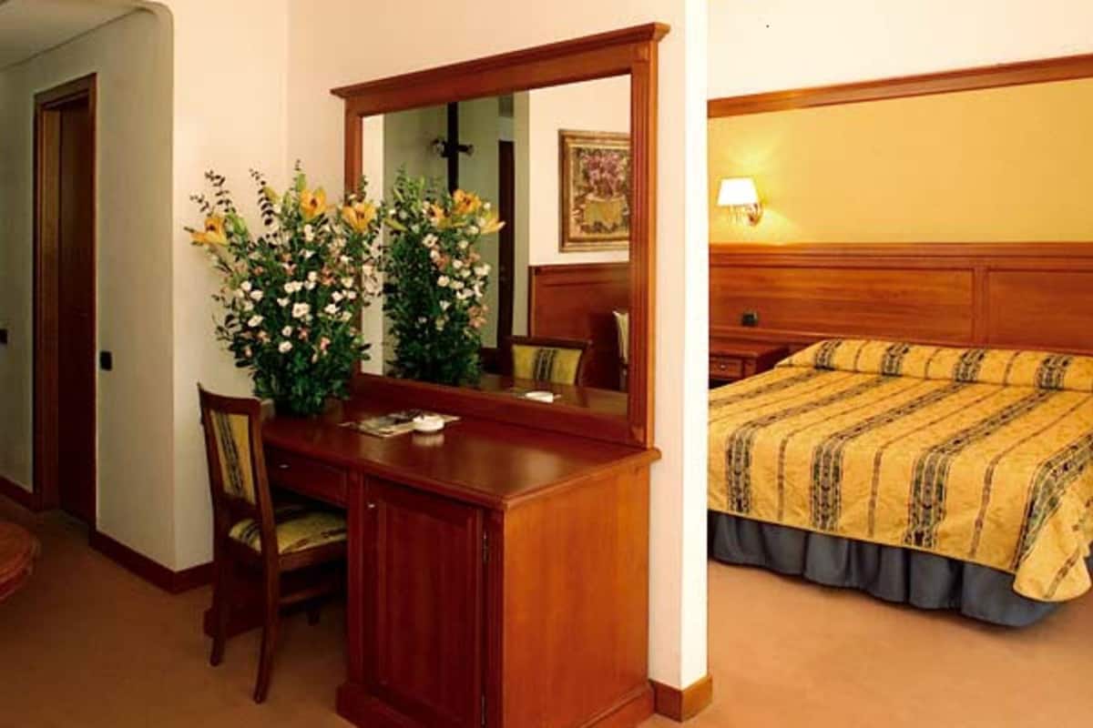 A hotel room with a wooden desk, a chair, a large mirror, a bed with a striped bedspread, and a nightstand with a lamp.