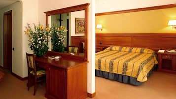 A hotel room with a wooden desk, a chair, a large mirror, a bed with a striped bedspread, and a nightstand with a lamp.