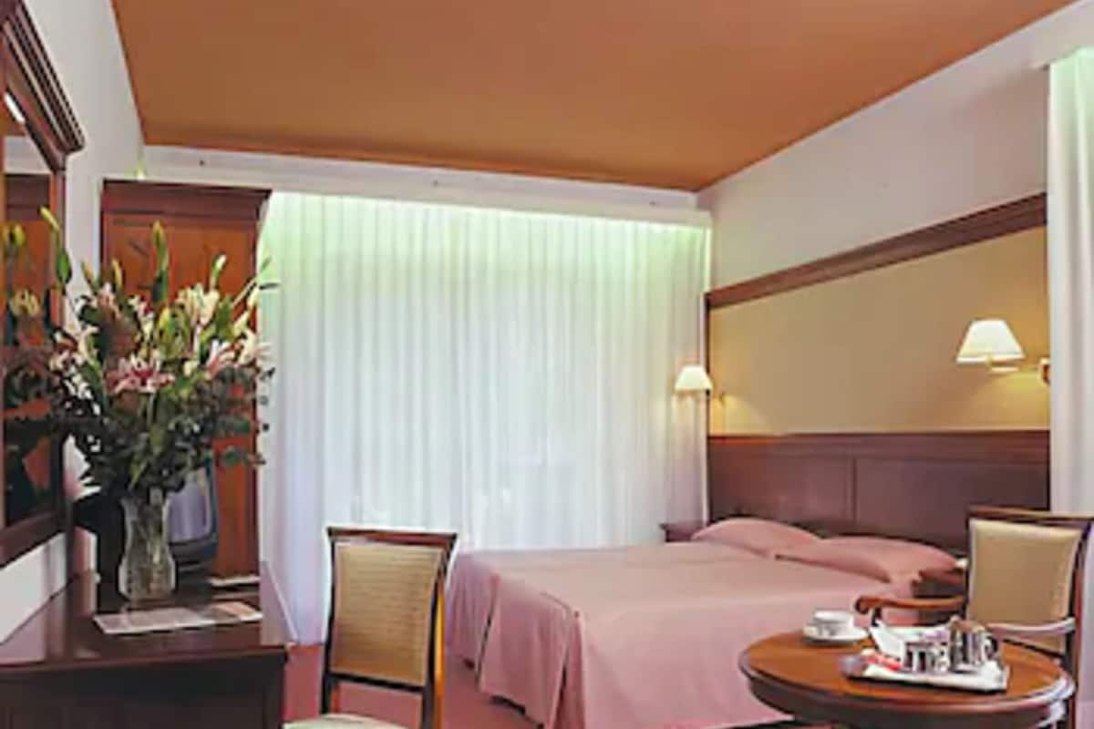 A hotel room with a bed, two chairs, a small table with a tray, a vase with flowers, and a window with curtains.