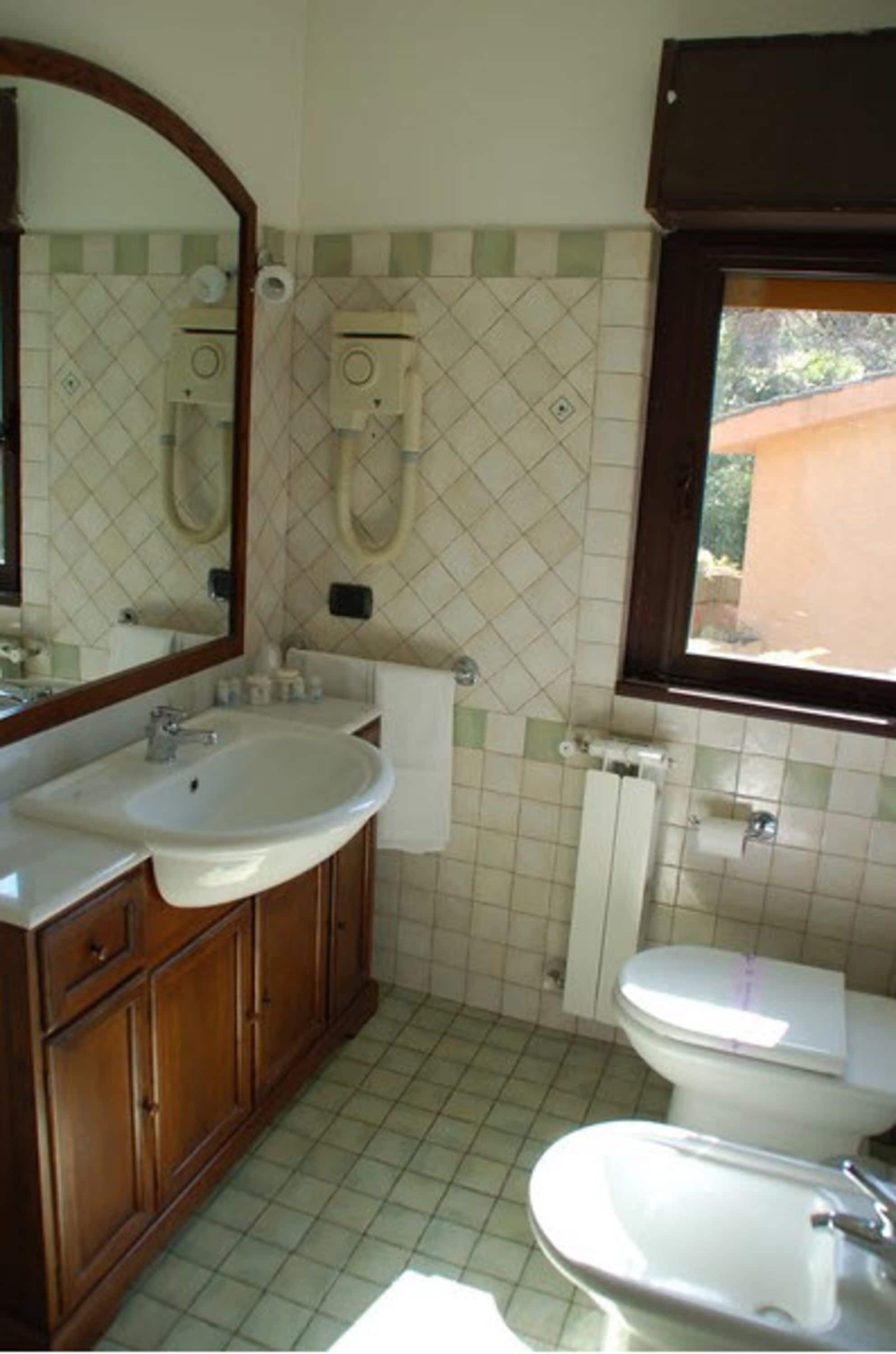 A bathroom with a sink, mirror, toilet, bidet, and a window.
