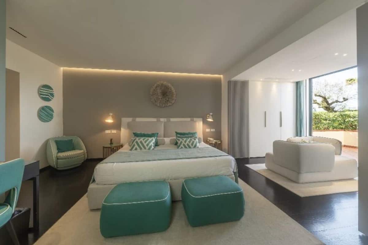 A modern bedroom with a large bed, two side chairs, a sofa, and a view of greenery outside.