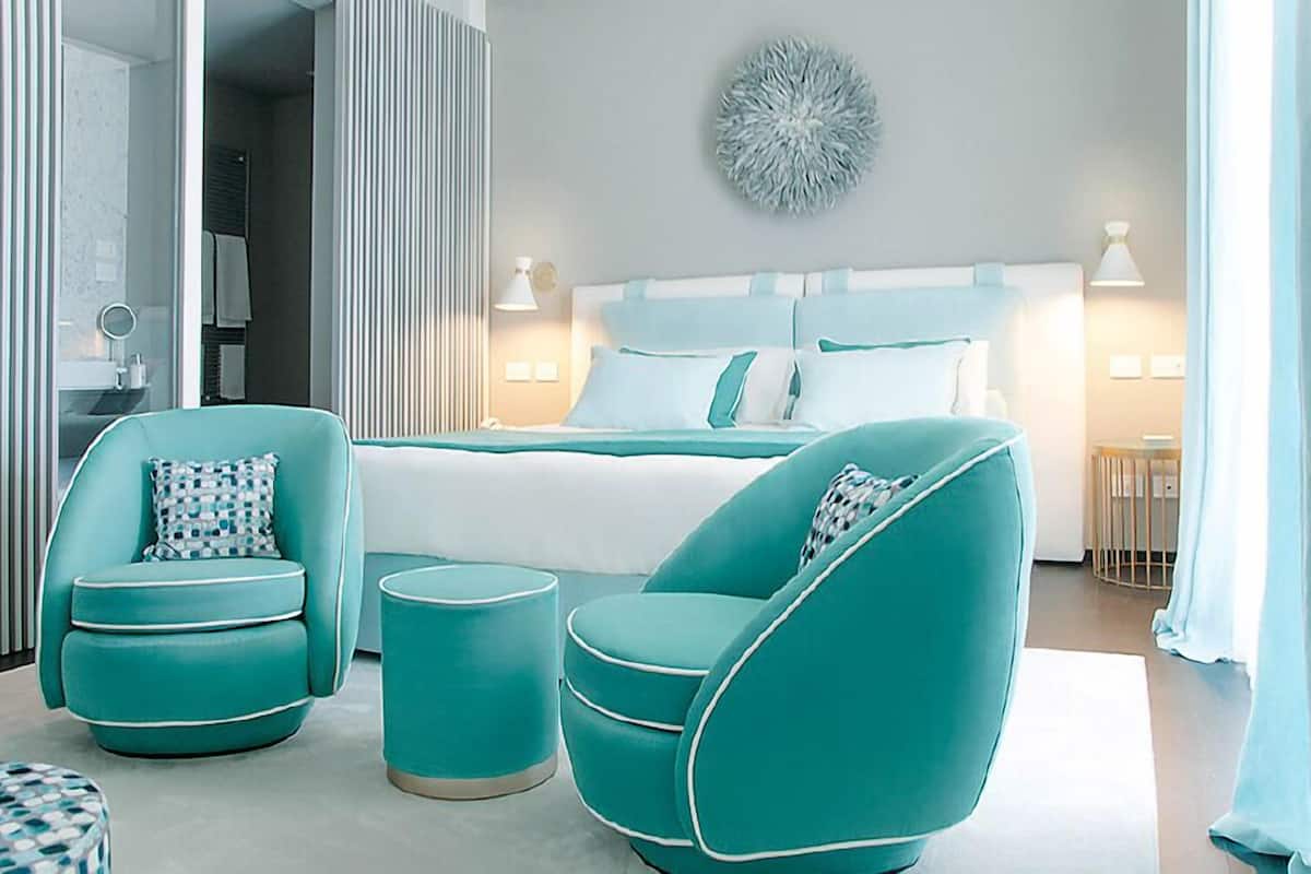 A modern bedroom with a large bed, two teal armchairs, and a round ottoman.
