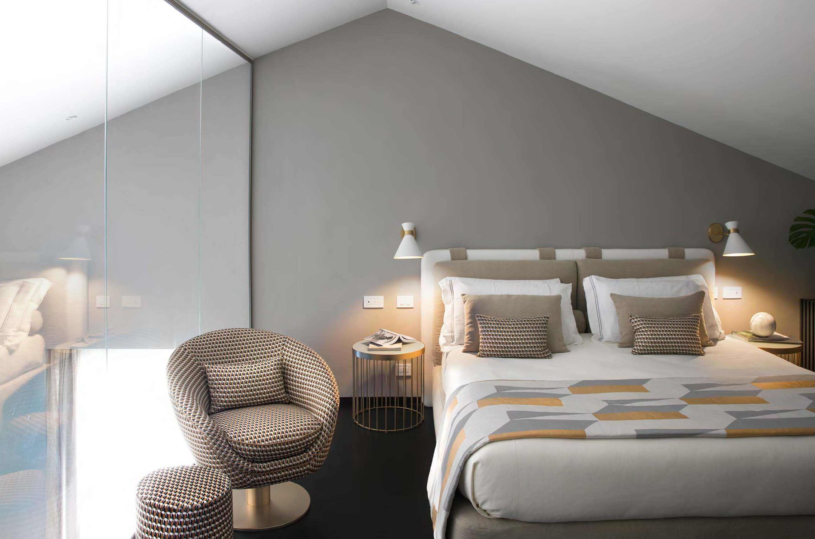Ein modernes Schlafzimmer mit Bett, Nachttischlampen, einem runden Beistelltisch, einem gemusterten Sessel und einem großen Fenster.