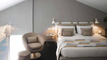 Ein modernes Schlafzimmer mit Bett, Nachttischlampen, einem runden Beistelltisch, einem gemusterten Sessel und einem großen Fenster.