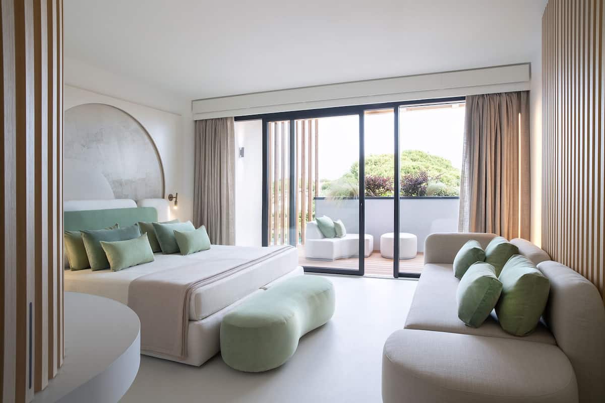 A modern hotel bedroom with a large bed, a sofa, and a balcony with a view of greenery.