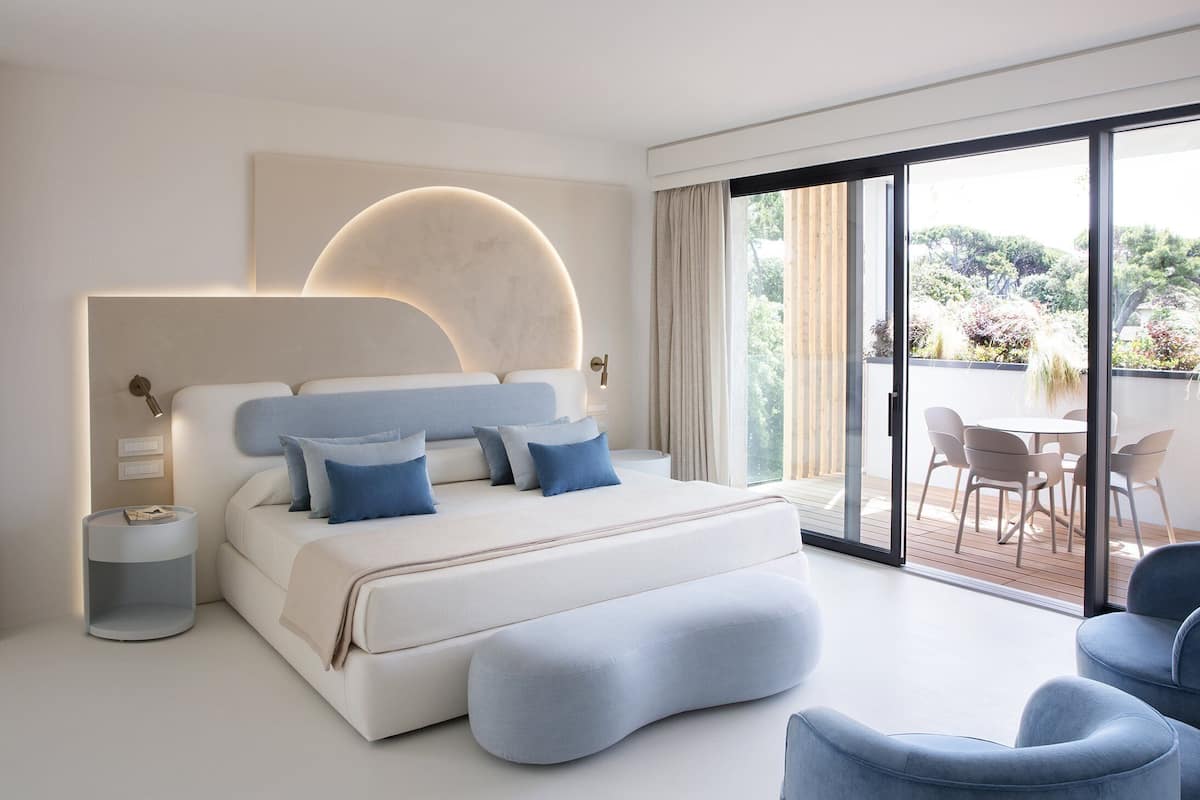 A modern bedroom with a large bed, a bedside table, and a view of an outdoor patio with chairs.