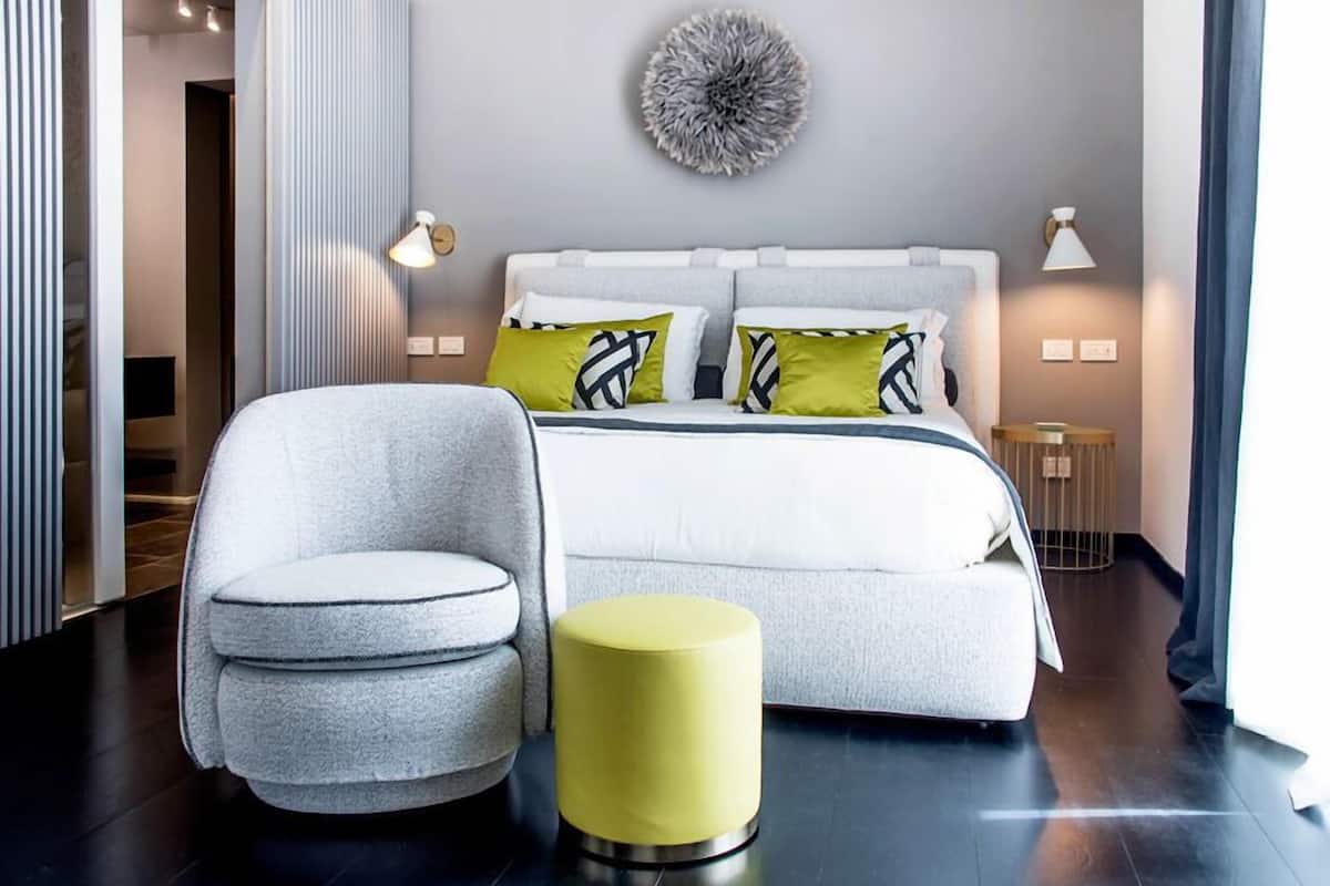 A modern bedroom with a bed, a grey armchair, a yellow ottoman, and a decorative wall piece.