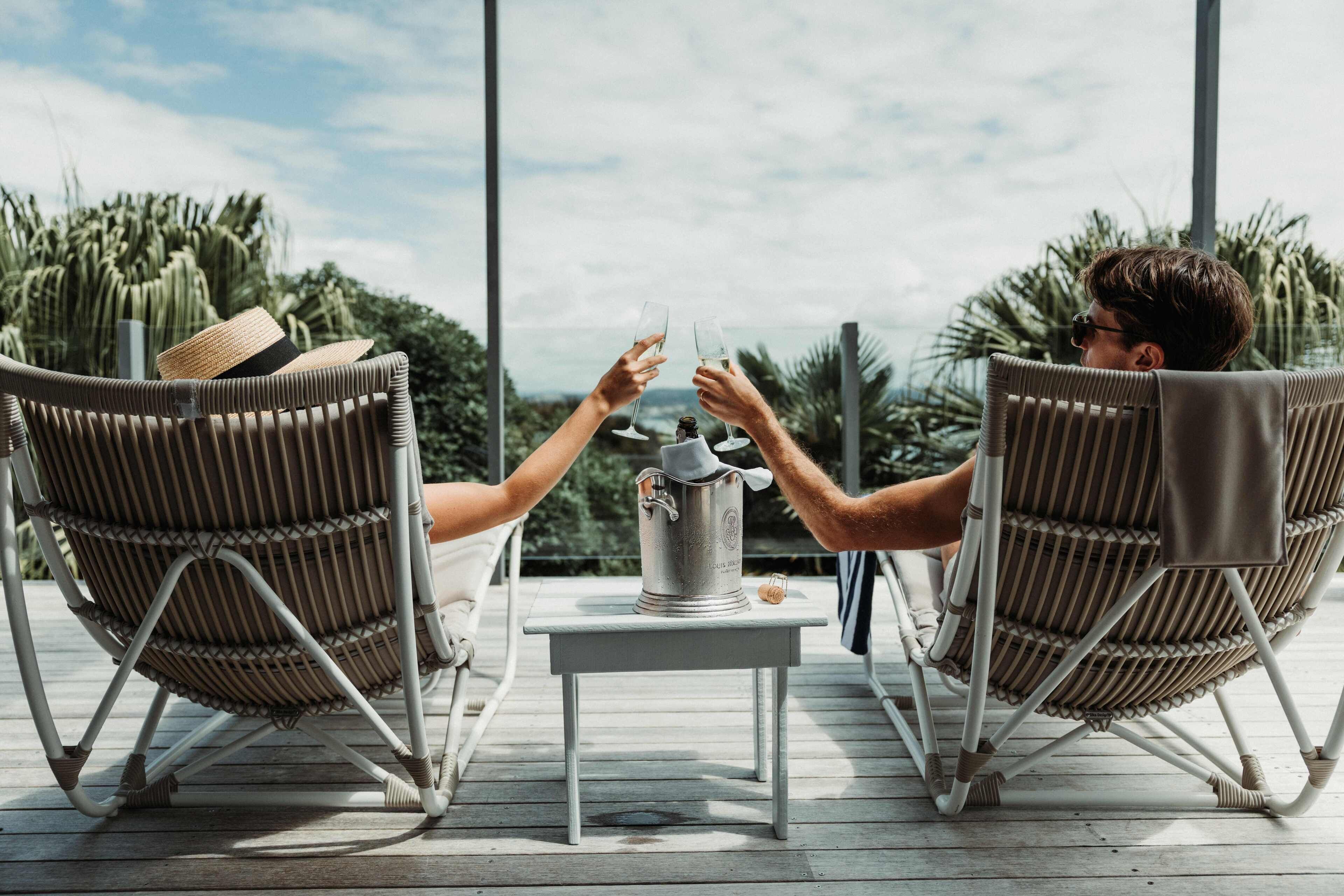 Two people toasting with champagne glasses on a wooden deck with wicker chairs, a small table, and a view of palm trees and mountains.