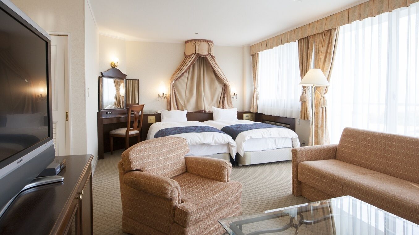 A hotel room with a bed, two bedside tables, a TV, a sofa, a chair, and a glass table.