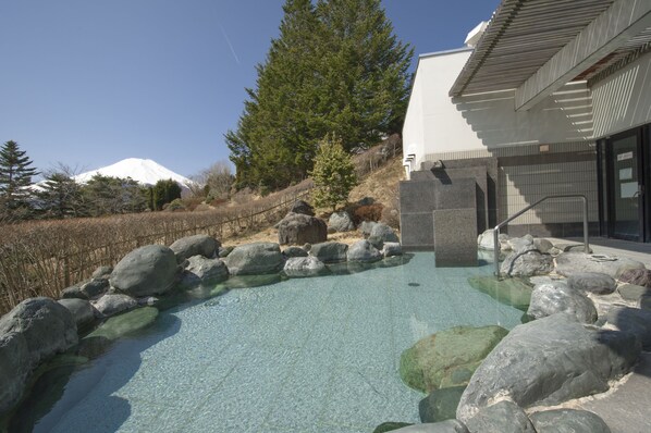 A clear outdoor pool with rocks, a modern building with a sloped roof, and a snow-capped mountain in the background. - Hotel Mt. Fuji (Yamanakako)