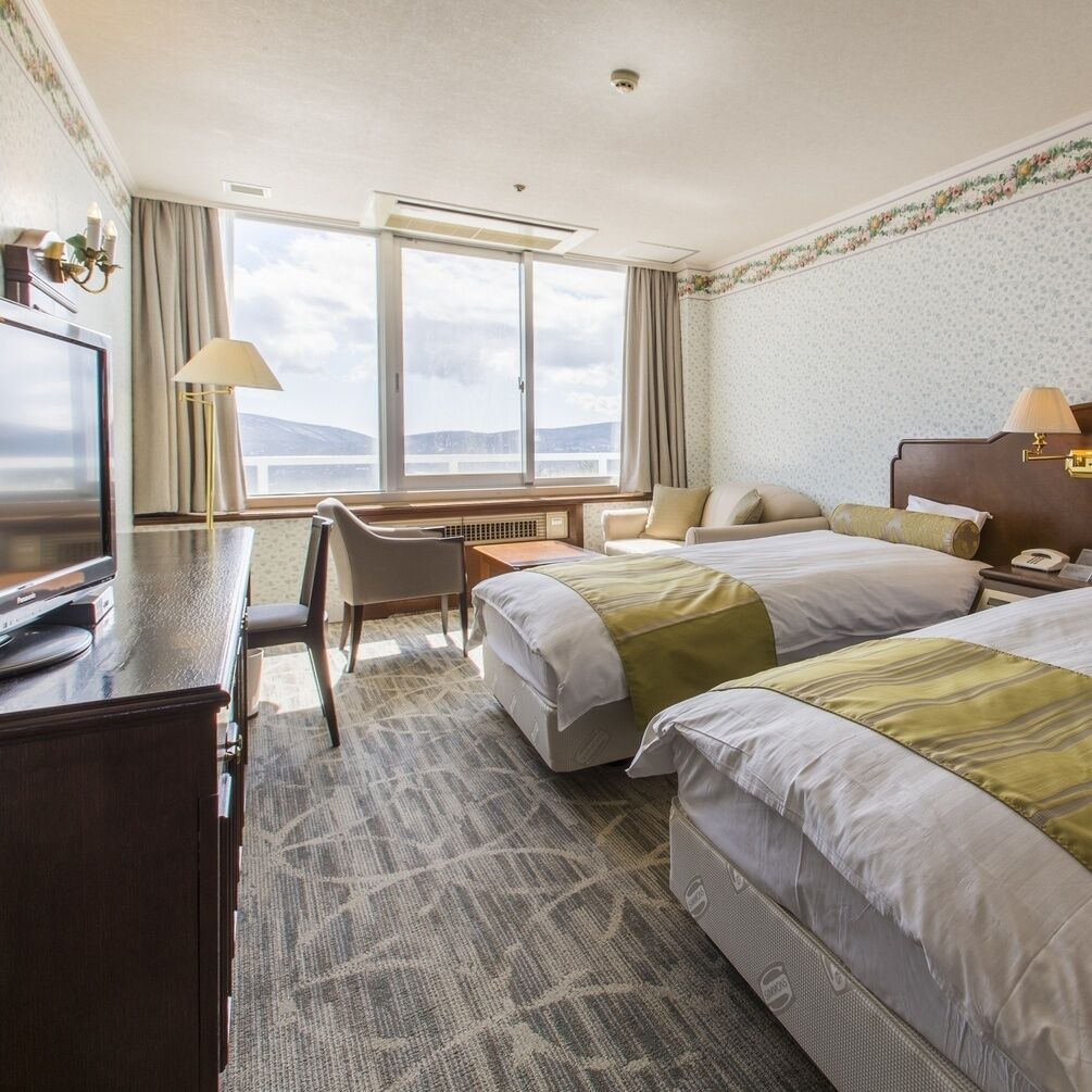 Quarto de hotel com duas camas, uma mesa de trabalho com cadeira, televisão e uma janela grande.