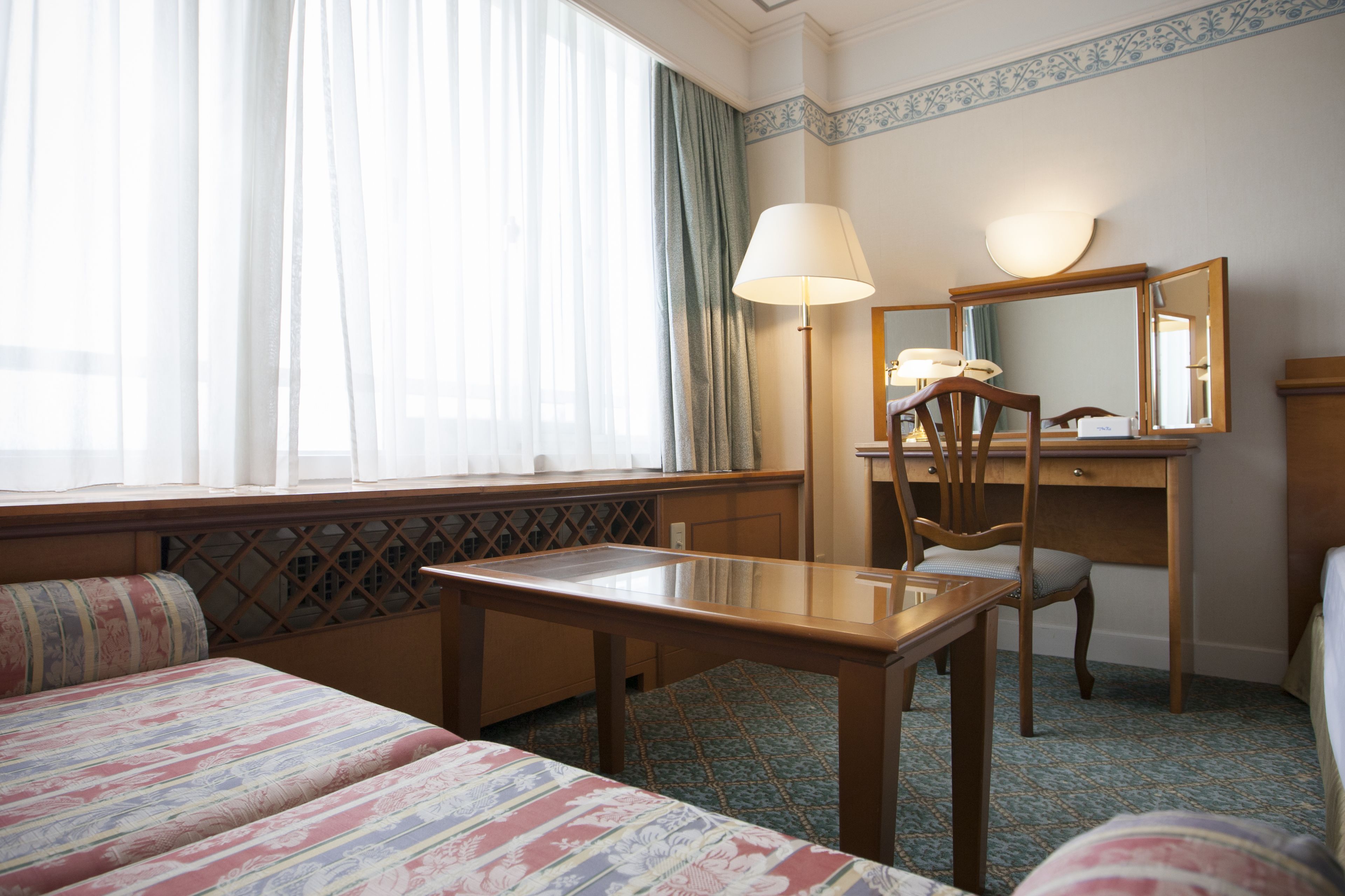 A hotel room with a bed, a desk, a chair, a lamp, and a large window with curtains.