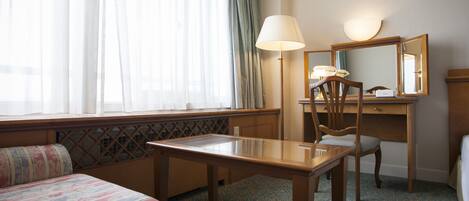 A hotel room with a bed, a desk, a chair, a lamp, and a large window with curtains.